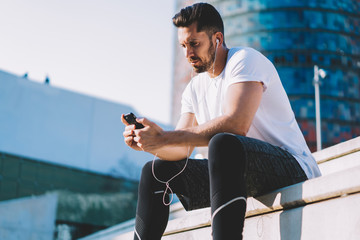 Confident muscular sportsman blogger using smartphone for share media in social networks having break on training, male athlete using earphones for listening music for active lifestyle workout