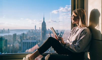 Side view of happy millennial digital nomad with modern touch pad looking in lookout window and dreaming about New York sightseeing, carefree hipster girl enjoying time in high rise building