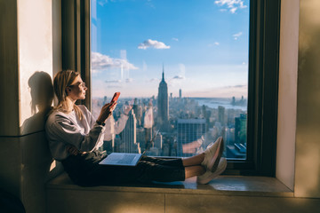 Youthful female freelancer holding modern smartphone device in hand and admiring beautiful scenery view of Manhattan sitting in high rise building with panoramic lookout, remote worker with laptop