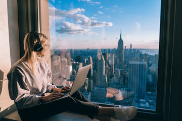 Millennial female freelancer in electronic headphones for noise cancellation listening positive music podcast and looking in panoramic window with breathtaking Manhattan view from high rise