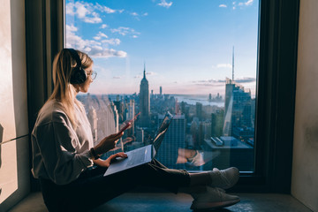 Digital nomad in bluetooth earphones enjoying leisure time for visiting high rise building with panoramic lookout on New York, millennial female with laptop and smartphone listening audio book - obrazy, fototapety, plakaty
