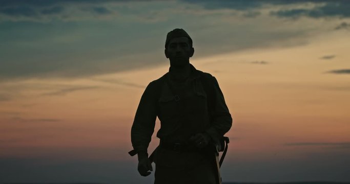 Soldier in uniform silhouetted against the sunset walking towards the camera across open countryside