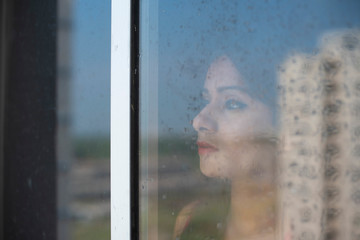 Blurred portrait of an young Indian brunette woman in red dress looking outside through a glass window with water droplets/dewdrops in home quarantine. Indian lifestyle and quarantine.