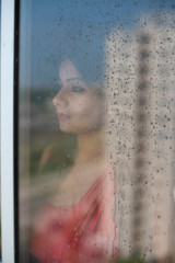 Blurred portrait of an young Indian brunette woman in red dress looking outside through a glass window with water droplets/dewdrops in home quarantine. Indian lifestyle and quarantine.