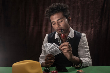 Portrait of young brunette Indian dark skinned man in vintage retro suit playing cards on a casino poker table in front of a textured copy space studio background. lifestyle and fashion.