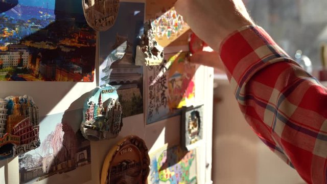 Man is fixing postcard on the fridge by adding new magnet to the collection.  Big collection from various locations is visible.
