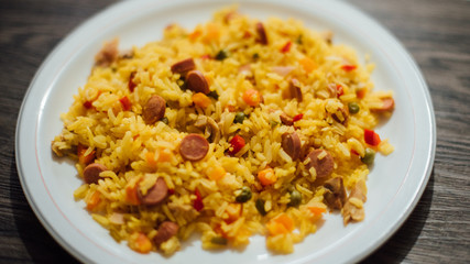 Rice, vegetables, onion, pea, carrot, peppers, chicken, sausage, ham in white plate on wooden table
