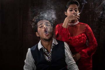 An young and attractive Indian brunette couple in vintage suits smoking in a casino poker table in...