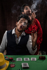 An young and attractive Indian brunette couple in vintage suits smoking in a casino poker table in brown textured copy space background. Gambling and addictive lifestyle.