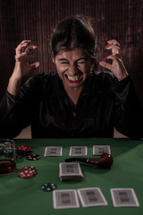 Young Indian Bengali brunette woman in western dress showing anger while playing cards on a casino poker table in brown textured copy space studio background. Indian lifestyle and fashion.