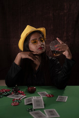 Young Indian Bengali brunette woman in western dress with yellow hat and whiskey playing cards on a casino poker table in brown textured copy space studio background. Indian lifestyle and fashion.