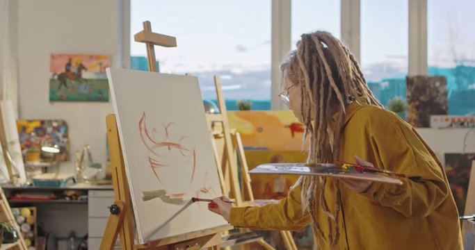 Stylish contemporary Caucasian female painter in glasses and with dreadlocks creating impressionism artwork on canvas in studio. Young man painting at easel in cozy workshop. Female artist.