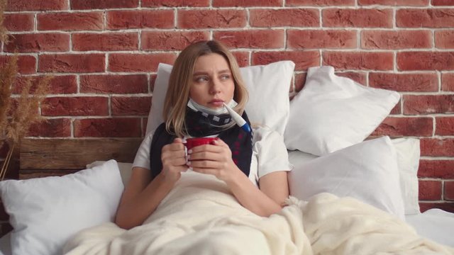 A sick girl with a scarf around her neck, lying in bed with a Cup of tea, measures and looks at the temperature taking a thermometer out of her mouth. A young sick woman in bed with a mug of tea