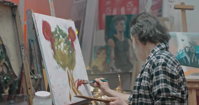 Caucasian middle-aged male painter drawing tree on canvas in impressionism style. Portrait of handsome happy man artist painting masterpiece with paints and brush, then turning to camera and smiling.