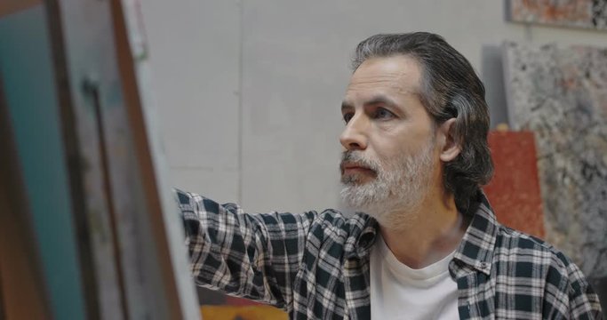 Close up of middle-aged Сaucasian handsome man with gray beard drawing at easel in cozy messy art studio. Artist working on canvas in workshop and creating new artwork. Male painter of fine art.