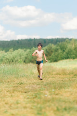 a boy in blue swimming trunks runs on a field of grass in the summer under the hot sun