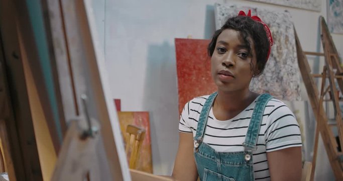 Portrait of young African American stylish female artist in painting picture on canvas and smiling cheerfully to camera. Talanted student at fine art course in academy. Pretty happy girl at easel.