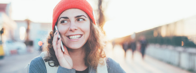 Smiling curly woman talking on the mobile phone on the street. Wearing red hat at sunset