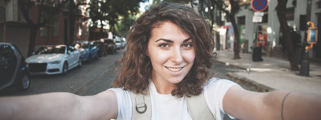 Cheerful curly woman taking a selfie photo on the street using her mobile phone, walking with backpack in the city