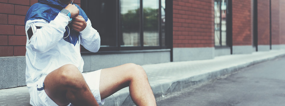 Young athlete in blue windrunner sitting on the street after good running session and resting