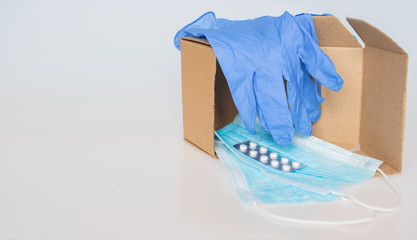 A single, moving cardboard box lies open on a white background, containing protective face masks and a package of pills, with blue latex medical gloves on top: humanitarian aid, medical concept, deliv