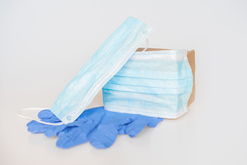 Single moving cardboard box in a face mask lies on a white background on a pair of blue doctor's protective gloves with another face mask on top: medical concept, humanitarian aid