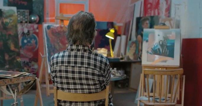 Rear of male Caucasian painter sitting on chair in front of his artworks and looking at them. Back view on man artist examining and evaluating pictures in workshop. Artistic atmosphere concept.