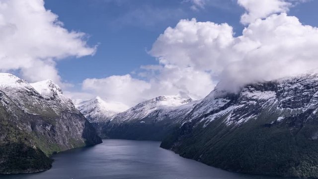 White clouds whirling above the calm blue water of the Geiranger fjord. Steep cliffsides covered with scarce vegetation. Fresh snow on the mountain tops.  Shadows dancing on the mountainsides.