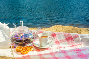 Glass teapot with fruit tea and cookies on a tablecloth on the background of a summer beach with sand and water