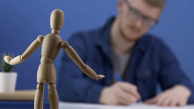 Designer artist paints a wooden gestalt figure. The work of the artist or designer. The creative process. Creative workspace in blue tones. A man with glasses is copying on paper a wooden man. Blur