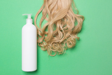 Lock of female blond hair and white bottle of lotion on green background. Top view
