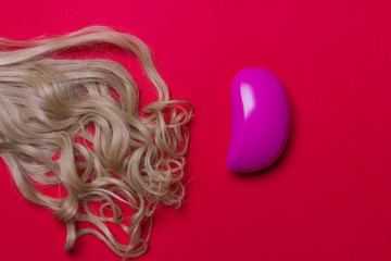 lock of woman blond hair and pink hairbrush on red colored paper background