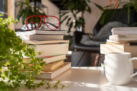 Inside a green house with many plants. A pile of different books on a with table with red glasses and a cup of coffee at sunset
