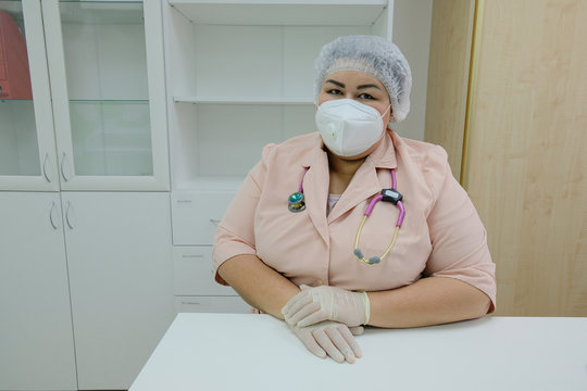 A Doctor In A Protective Mask And Gloves Sits At A Desk And Takes A Reception During The Coronavirus Pandemic.