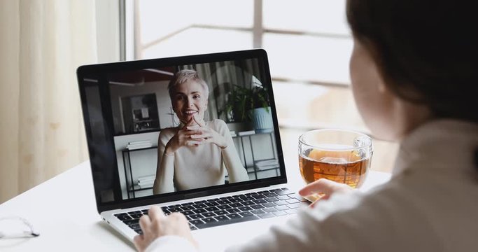 Two young business women distance workers talk use videoconference chat on laptop. Female online teacher, coach, manager video calling in remote home office conference on screen. Over shoulder view