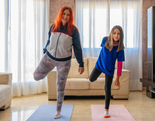Fototapeta na wymiar Mother and daughter dressed in sportswear exercising between white sofas in the living room at home