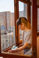 Portrait of a Caucasian boy with medical mask stands at the window at home looking out the window. Misses you at home. Quarantine protection against coronavirus. Home insulation