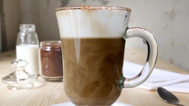 Swirls of milk mixed with dark coffee in cup. Cappuccino or latte in tall glass with handle. Two-layer drink with froth milk. Close-up side view, white tray, wooden table, granulated coffee in jar