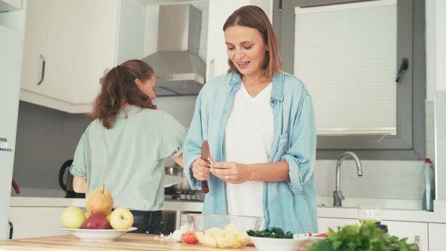 Two young pretty women singing and dancing while cooking in the bright kitchen. Friends are having fun while cooking together.