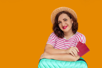 Portrait of a beautiful smiling woman with dark curly hair in a boater hat on a yellow background. a woman holds a passport in her hands and leans on a suitcase. awaiting travel