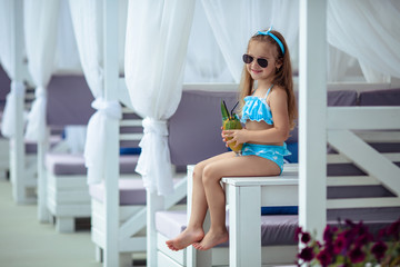 Little beautiful girl of 5 years with dark hair in a blue polka dot swimsuit and in sunglasses drinks a cocktail on a summer vacation by the pool.