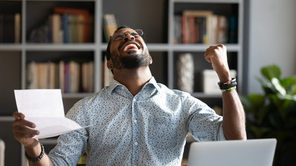 Overjoyed young mixed race man in glasses holding paper in hands, celebrating unbelievable triumph,...