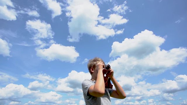 Closeup view video of young white kid holding old retro photo camera in hands taking pictures of beautiful sunny summer landscape. Kid isolated at clear blue sky backround.