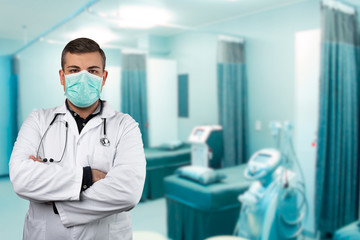 Fototapeta na wymiar Caucasian man posing as a medical doctor in white overall or scrub with protective face mask and stethoscope around a neck. Hospital background.