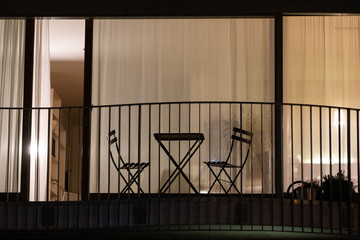 Tisch und Stühle stehen auf einem Balkon vor dem abendlich erleuchteten Wohnungsfenster.