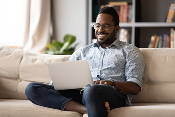 Smiling young african american man in eyewear relaxing on couch, looking at laptop screen. Happy multiracial guy web surfing internet, chatting with friends in social network, watching movie at home.