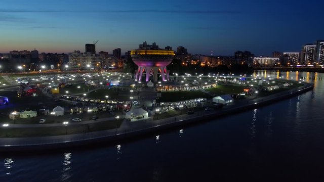 Aerial forward festive night scenic city landscape Kazan illumination Unique cauldron steel boiler national style modern architecture on the island river bank. Travel attraction. People walk promenade
