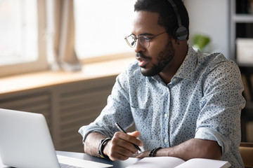 Head shot focused young african american man in glasses wearing wireless headphones, looking at laptop screen, watching educational lecture seminar webinar online, studying remotely, writing notes. - Powered by Adobe