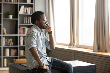 Involved in interested mobile conversation happy handsome biracial guy sitting on desk in office or home. Smiling young african american businessman talking on smartphone with clients partners.