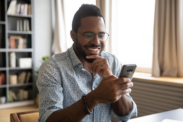 Head shot happy young african american man in eyeglasses looking in phone screen. Smiling...